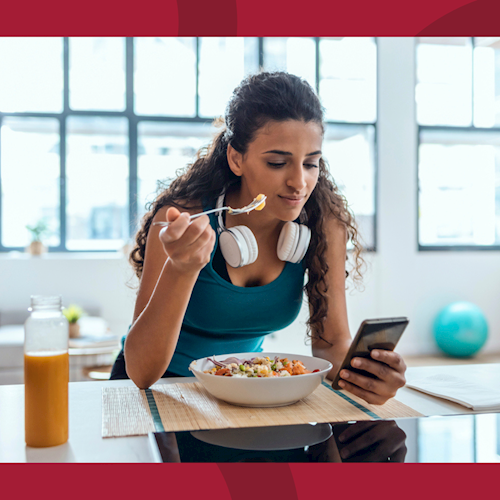 Woman with headphones around her neck is eating cereal while looking at her phone.